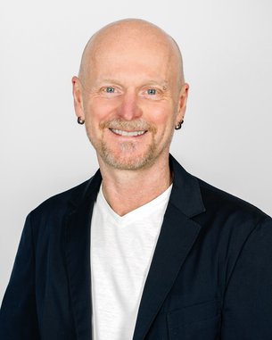 Mann mit Glatze und Ohrstecker, trägt einen schwarzen Blazer über einem weißen T-Shirt, lächelt freundlich vor neutralem Hintergrund.